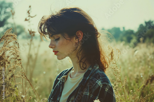 A young woman with brown hair and red lipstick, wearing a plaid shirt, stands in a field of tall grass, looking down and away from the camera photo