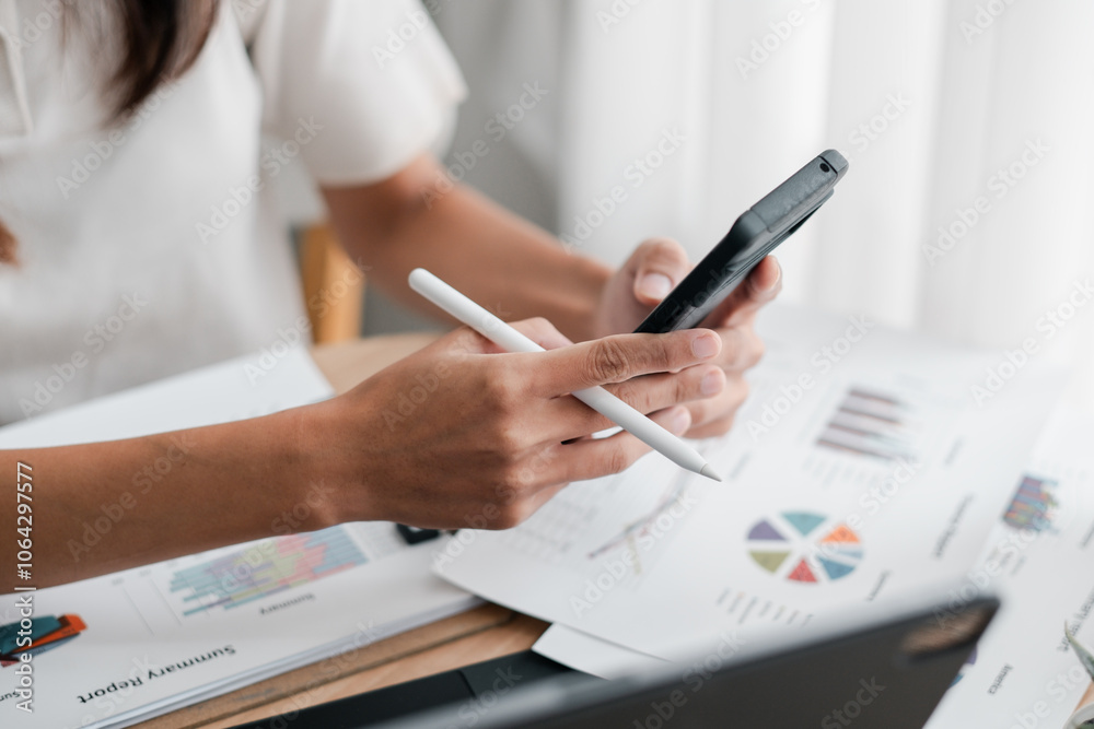 Close-up of hands using a smartphone and stylus to review financial charts and graphs on paper in a bright office. Close-up of hands using a smartphone and stylus to review financial charts and graphs on paper in a bright office.