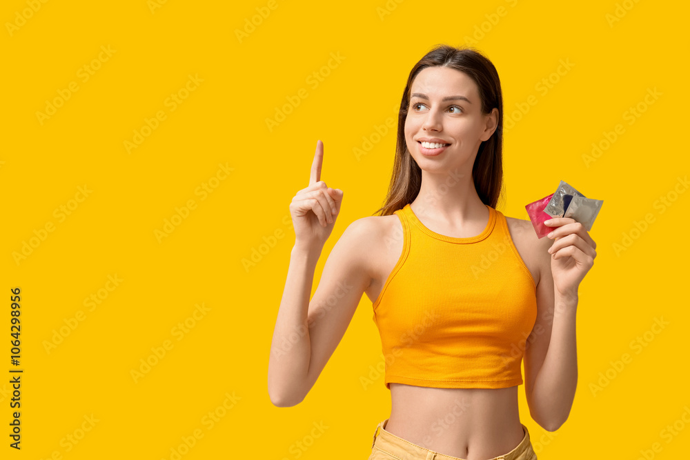 Beautiful young woman with condoms pointing at something on yellow background Beautiful young woman with condoms pointing at something on yellow background