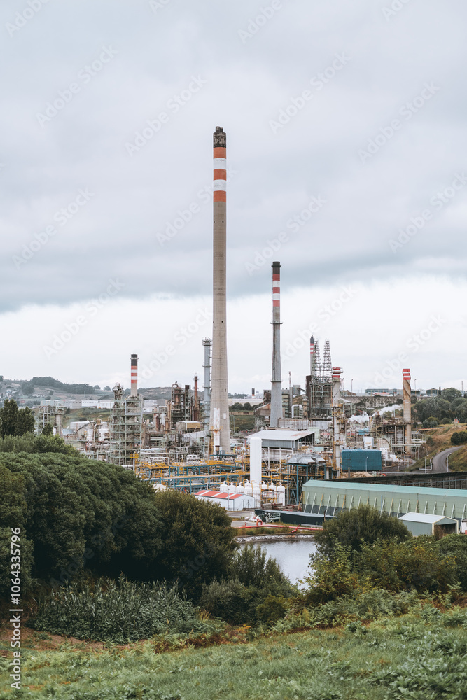 Obraz premium A large industrial refinery complex with tall chimneys emitting light smoke, surrounded by lush green trees and hills under a cloudy sky. Detailed infrastructure visible