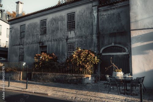 Old stone building facade with weathered walls and barred windows, adjacent to a small outdoor dining area with a table, chairs, and wine barrel planter. Golden hour lighting creates warm shadows