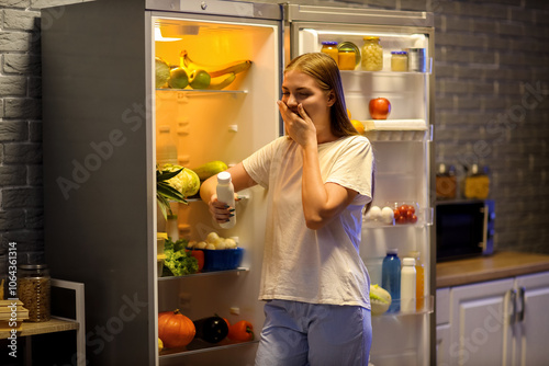 Sleepy young woman with yoghurt near open fridge in kitchen at night