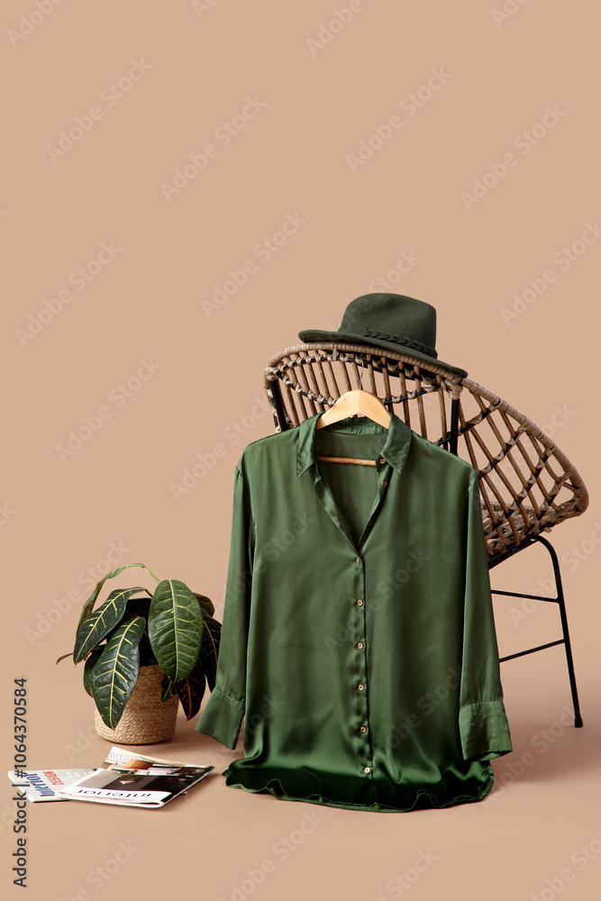 Obraz premium Hanger with stylish female blouse hanging on armchair, hat, houseplant and magazines against color background