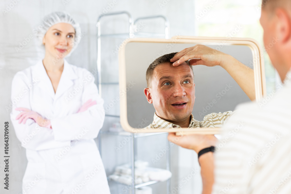 Obraz premium Satisfied middle-aged man patient holding big mirror sitting on clinical chair in aesthetic clinic