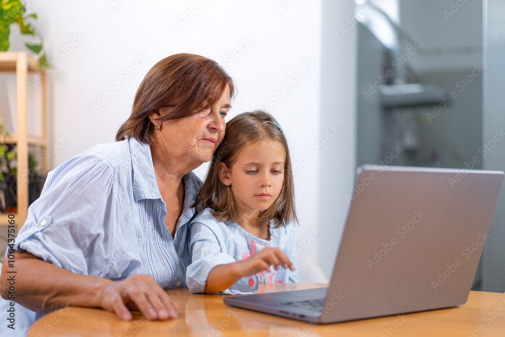 Granddaughter and grandmother sit together at table using laptop, sharing moment of learning and connection in comfort of their home Granddaughter and grandmother sit together at table using laptop, sharing moment of learning and connection in comfort of their home