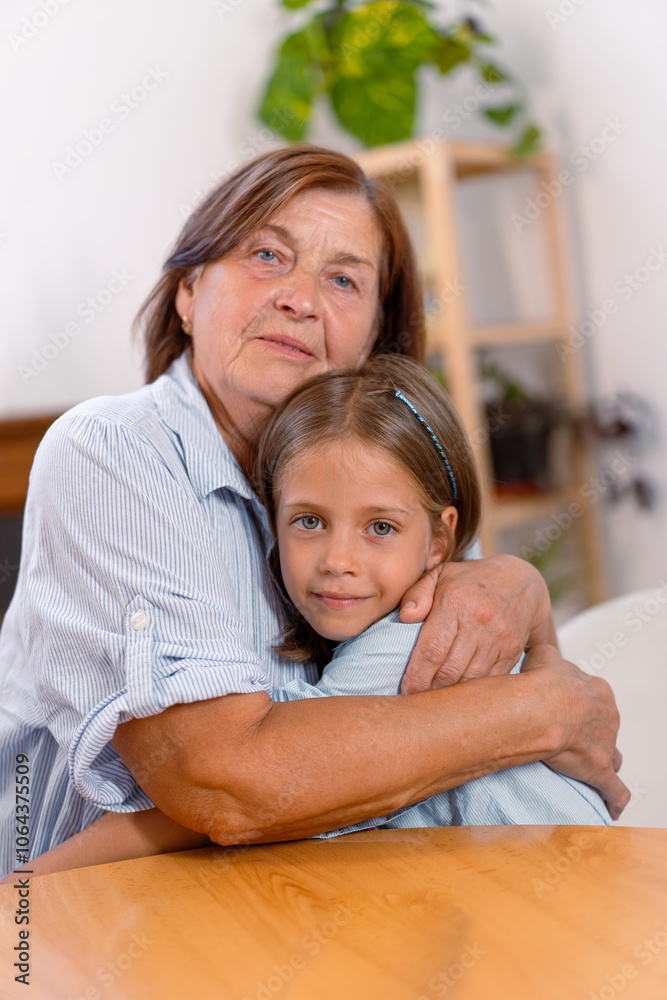 Loving grandmother and granddaughter share warm embrace at home, their faces filled with joy as they look at camera, capturing heartfelt moment of connection across generations Loving grandmother and granddaughter share warm embrace at home, their faces filled with joy as they look at camera, capturing heartfelt moment of connection across generations
