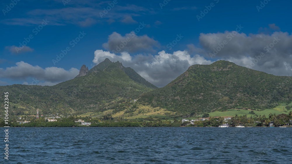 The picturesque landscape of Mauritius. Green mountains against blue sky and clouds. On the coast of the blue ocean, buildings are visible among the trees. The boats are moored near the shore.