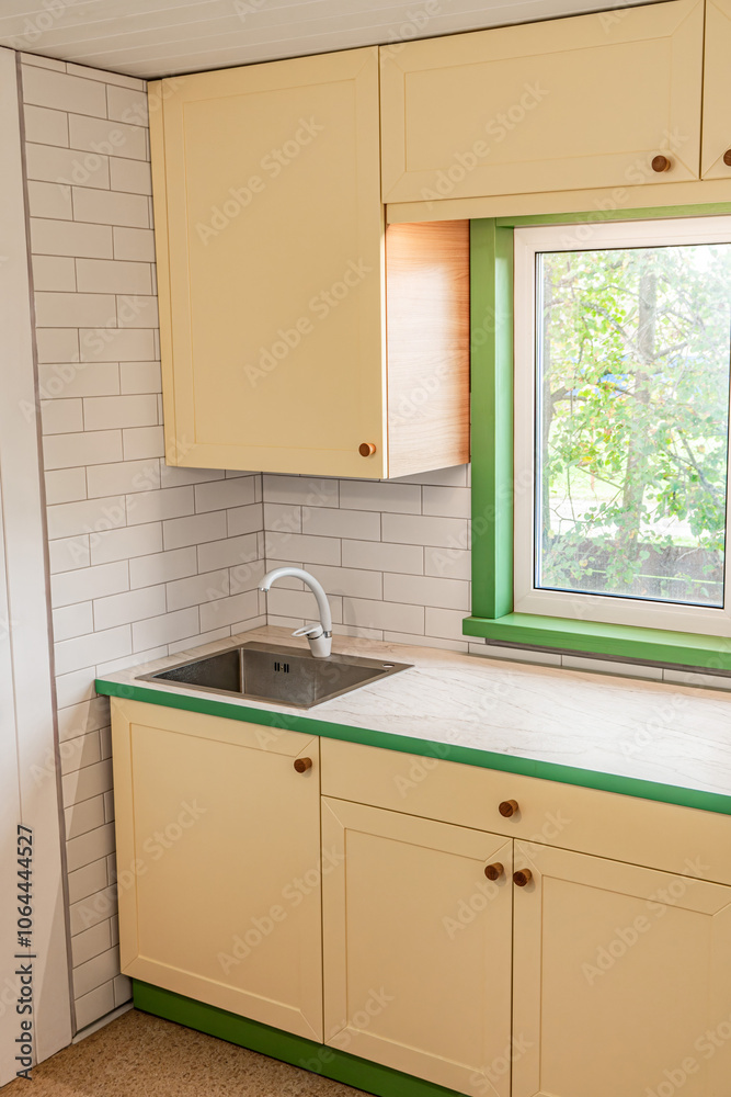 Fragment of kitchen with yellow cabinets with wooden handles features a plastic countertop with a marble-like finish, and upper cabinets installed around the window Fragment of kitchen with yellow cabinets with wooden handles features a plastic countertop with a marble-like finish, and upper cabinets installed around the window