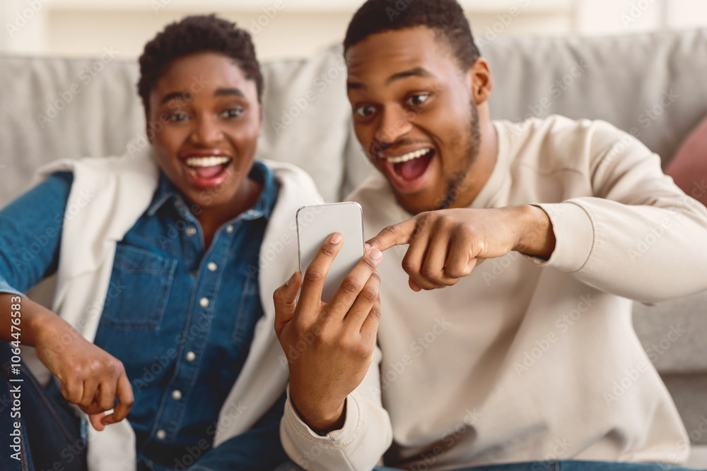 Wow. Portrait of overjoyed young black couple sitting on the floor, using smartphone. Excited guy showing his phone and pointing at screen, sharing amazing content, selective focus on device