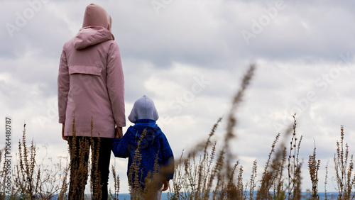 A mother and her young child enjoy together a peaceful walk through a meadow, holding hands as they observe the distant sky and surrounding nature. Family bonding. Weekend activities.