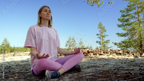 A young woman enjoys a moment of stillness as she meditates while hiking in a tranquil forest. The clear blue sky and surrounding trees provide a calming backdrop for her practice.