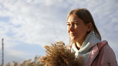 A young woman embraces her natural beauty while enjoying a peaceful moment outdoors. She holds a bouquet of grasses, radiating freedom and tranquility against the stunning sky.