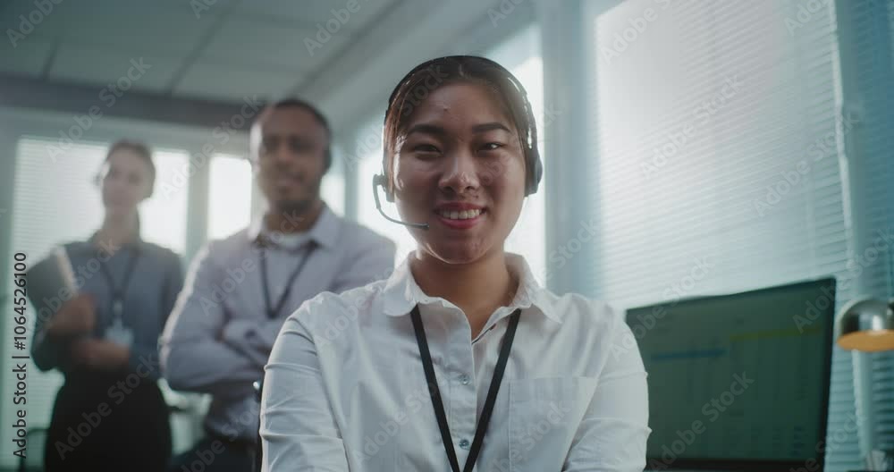Call center office: Close up portrait of friendly Asian female technical customer support specialist wearing headset smiling, looking at camera. Team of hotline operators standing in the background.