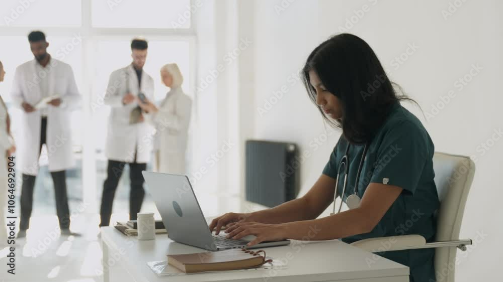 Side view of indian female doctor using portable computer while sitting at office desk in health center interior. Efficient young general practitioner checking patient records via gadget laptop Side view of indian female doctor using portable computer while sitting at office desk in health center interior. Efficient young general practitioner checking patient records via gadget laptop