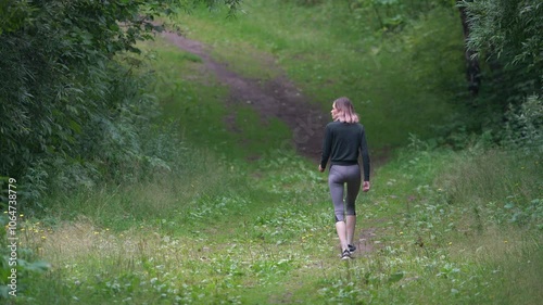 Sporty woman during summer sports training walking along footpath in park. Unrecognizable fit model dressed in sportswear - tracksuit jacket, cropped yoga leggings, sports shoe. Full length, rear view