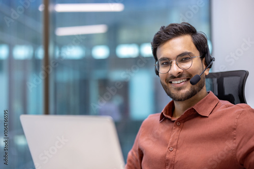 Hispanic businessman wearing headset working on laptop in office setting. Confident smile, brown shirt, glasses, engaging with professionalism. Bright, modern background. Business and communication.