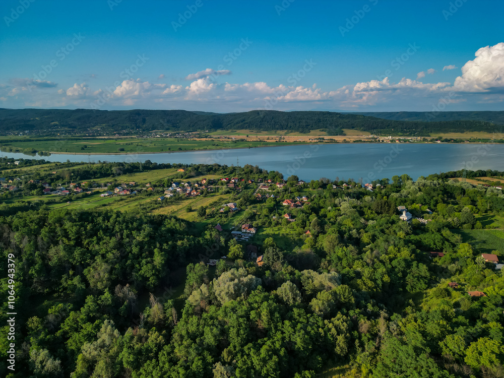 Fototapeta premium Panorama with a storage dam captur with a drone in the countryside. A calm river that comes from the mountains among the green forests and flows into the lake at the edge of a village in Romania