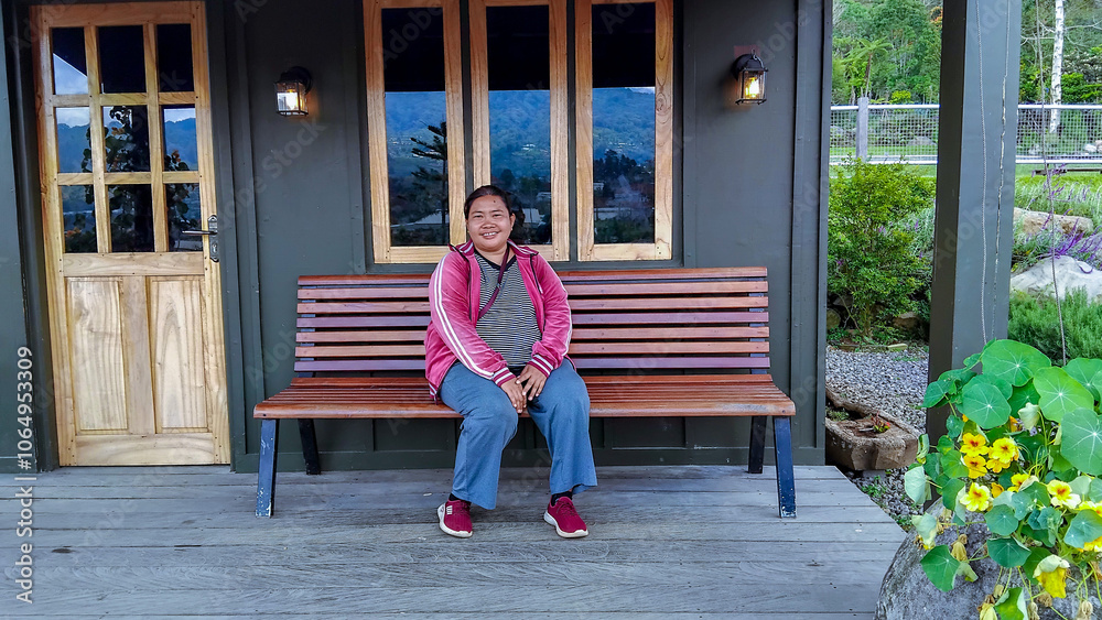 A woman visitor sits happily on a wooden bench by a small garden house in a scenic mountain area, surrounded by lush plants and flowers, enjoying the peaceful atmosphere
