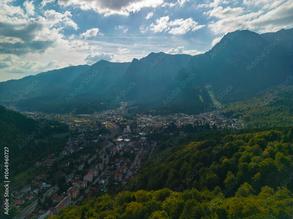 Fototapeta premium A town at the base of the mountain in the autumn season. Astonishing aerial view with the rocky mountains filmed in cinematic style. Romanian landscape above Busteni city in a sunny day with blue sky