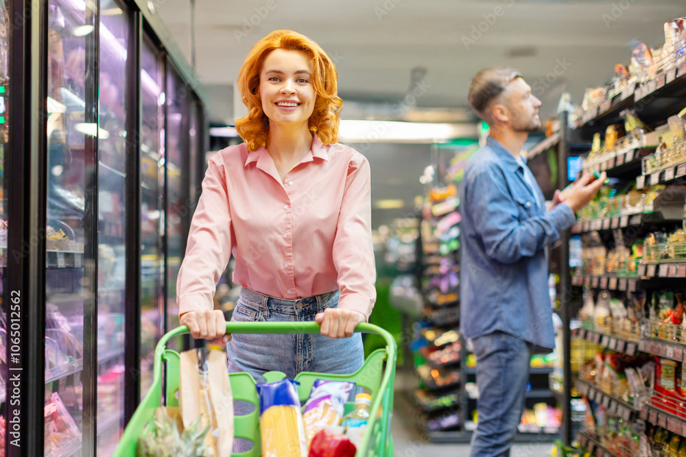 Obraz premium Cheerful lady doing shopping, buying products in supermarket, pushing trolley cart and smiling at camera