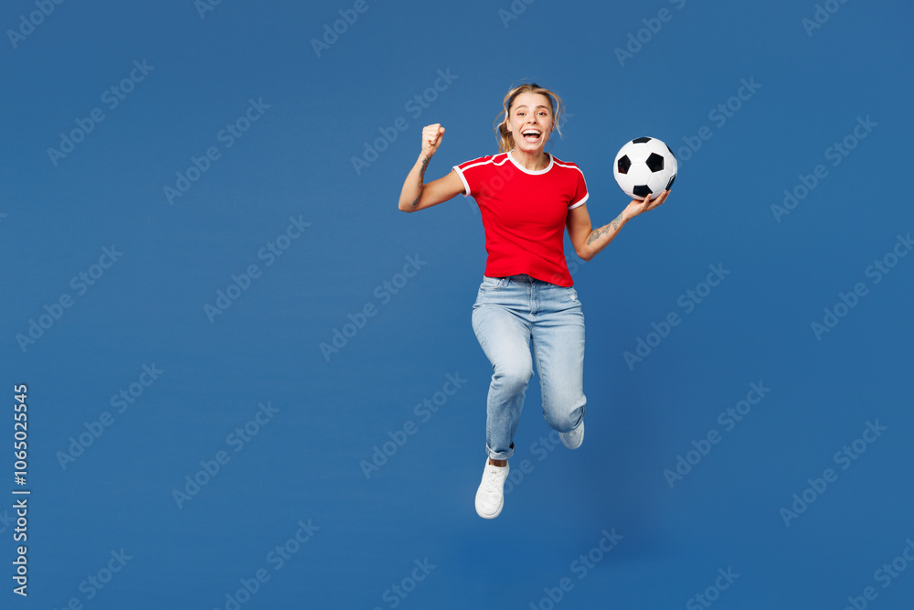Full body young woman fan in red t-shirt cheer up support football sport team hold in hand soccer ball watch tv live stream jump high do winner gesture clench fist isolated on plain blue background