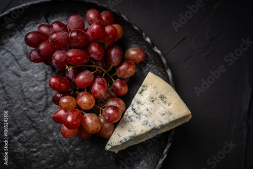 Blue blue cheese Garganzola or Dor Blue with a bunch of red grapes and a yellow pear on a black mica board on a dark background. Space for text. Top view photo