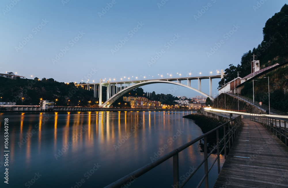 Obraz premium Arrabida bridge with car light trails reflecting on douro river at blue hour with illuminated cityscape and wooden walkway in Porto, Portugal