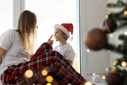 family happily spends time together on Christmas morning. child and mom sit on windowsill near window and tree with a glowing garland in foreground. positive mood, Christmas Eve at home