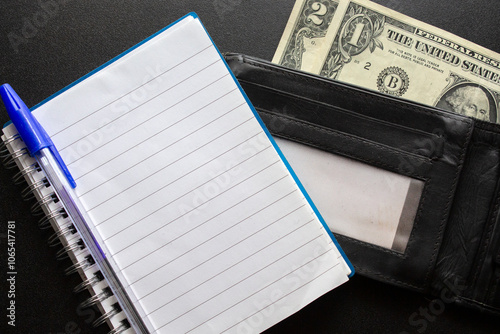 notebook with a blue pen on , next to a black leather wallet with US dollar bills. 