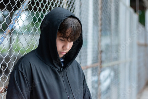 Asian youngster wearing black hoody and standing beside a metal fence in a local detention center, looking sad and wanting his freedom, broken home, lonliness, depression of young people concept. photo