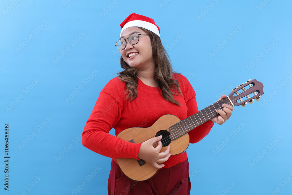 Obraz premium Young woman in Santa hat playing acoustic guitar on blue background. Christmas music