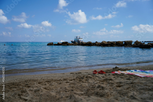 Red Slippers and Colorful Towel on Sandy Beach with Blue Sea and Boat in the Background
