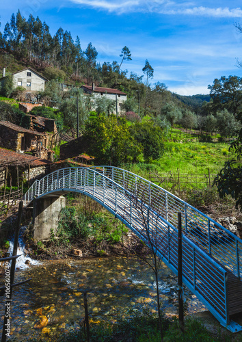 rural scene featuring a metal footbridge that spans a gently flowing stream, connecting lush green fields on both sides