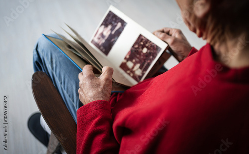 Картината върху платно Lonely old man in red sweater sitting on chair holding photo album looking at ph