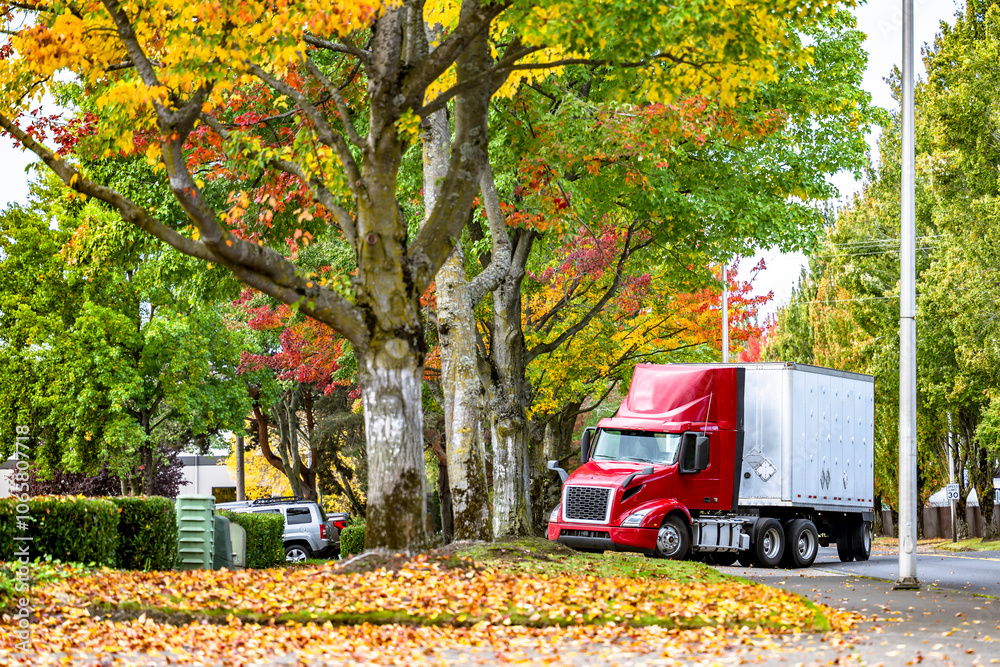 Obraz premium Local delivery red day cab big rig semi truck with roof spoiler and dry van semi trailer turning to warehouse parking lot from autumn road with yellow and red maples with partially fallen leaves