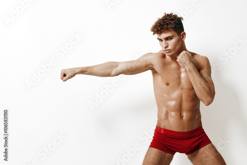 Muscular young man practicing boxing stance in red shorts, demonstrating focus and determination against a white background. Sports and fitness concept. photo