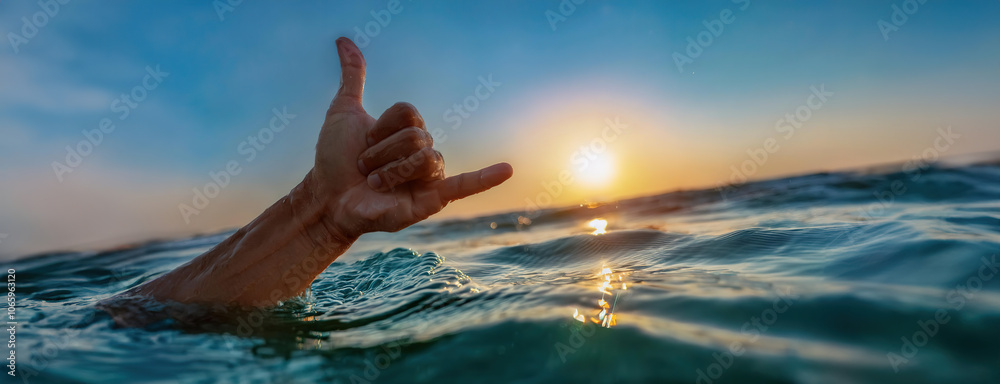A hand making the shaka gesture while floating in the ocean at sunset.