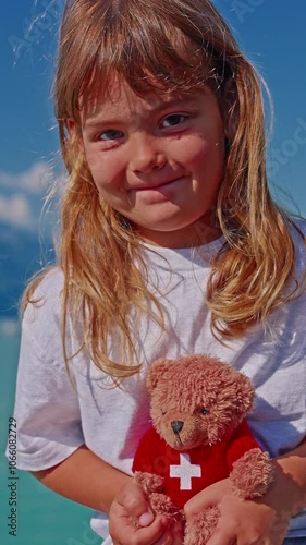 Little girl holding a small soft teddy bear with the image of the Swiss flag as a symbol of Switzerland against the background of the beautiful lake Interlaken in the city of Brienz photo