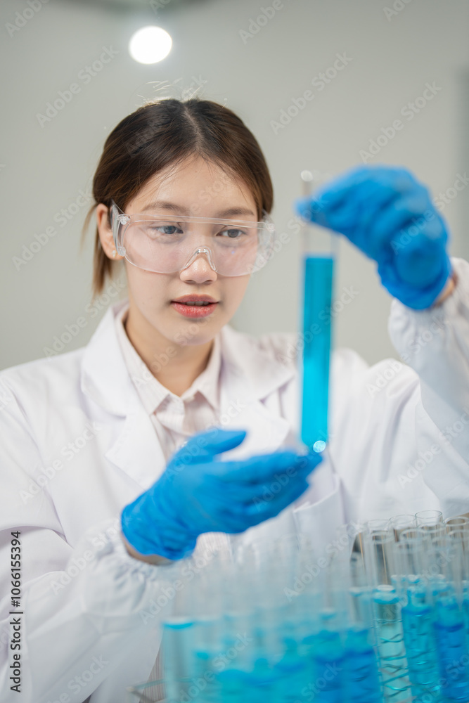 Fototapeta premium Young scientist wearing lab coat and gloves carefully examining test tube filled with blue liquid, conducting research or experiment in modern laboratory setting