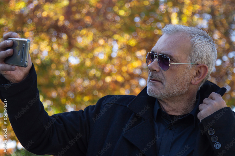 Mature man using mobile phone in the park in autumn for taking a selfie or live video Mature man using mobile phone in the park in autumn for taking a selfie or live video