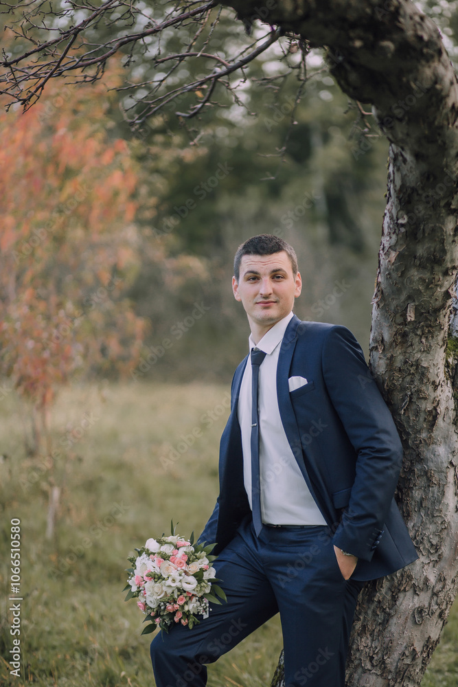 A man dressed in a suit and tie stands next to a tree, holding a bouquet of flowers. He is posing for a photo, possibly for a wedding or other special occasion A man dressed in a suit and tie stands next to a tree, holding a bouquet of flowers. He is posing for a photo, possibly for a wedding or other special occasion