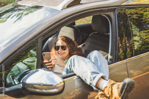 Happy young woman driver relaxing with smartphone in hands lying in car