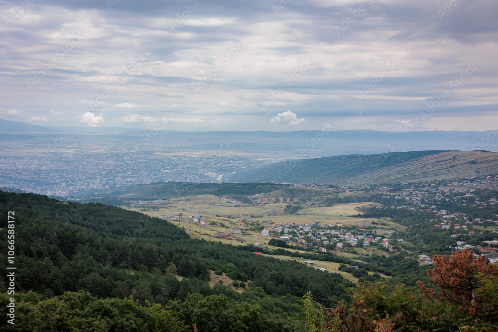 Obraz premium Summer mountain view of Tbilisi, Georgia. Soft clouds cast gentle light over the city and its green landscape, with lush trees in the foreground, creating a calm, scenic atmosphere