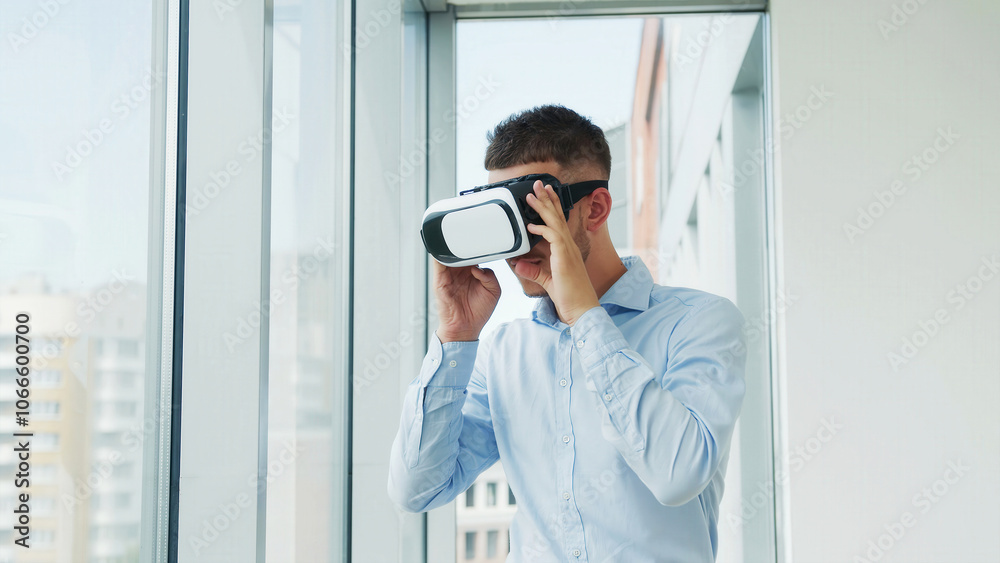 Man Using Virtual Reality Headset in Modern Office. A young man in a light blue shirt explores virtual reality using a VR headset in a bright, modern office with large windows. Man Using Virtual Reality Headset in Modern Office. A young man in a light blue shirt explores virtual reality using a VR headset in a bright, modern office with large windows.