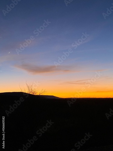 atardecer ladera de la montaña vista desde la carretera, silueta con poca vegetación, línea naranja franja de sol, cielo azul añil casi violeta decorado con pocas nubes y una pequeña luna al fondo photo