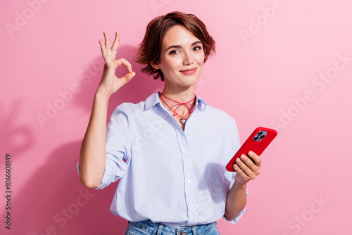 Photo of attractive woman with accessories dressed blue shirt hold smartphone showing okey approve isolated on pink color background