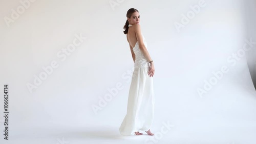 Young fashion model posing in long stylish Flowing white dress against isolated white background in studio. Woman beauty and grace