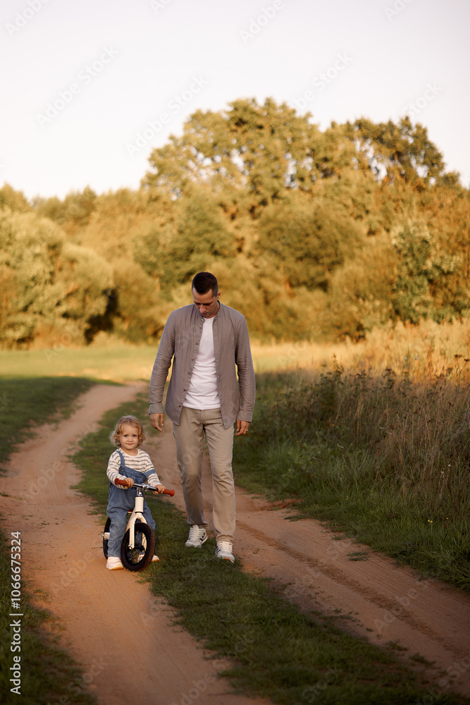Obraz premium Father teaches a young child to ride a balance bike in the park in the summer. The concept of fatherhood and family weekend outings.