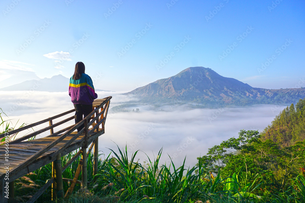Fototapeta premium Woman standing alone from behind on a wooden bridge, looking at the view of clear blue sky over mountains with fog in the morning. Inspirational motivational background. Mount Batur, Kintamani, Bali