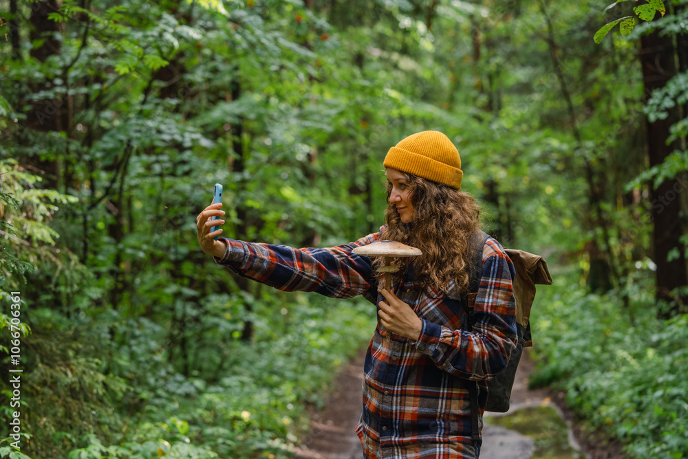 Fototapeta premium Young biologist or hiker woman wearing yellow beanie and plaid shirt taking a selfie with a mushroom she found in a green forest during a summer hike